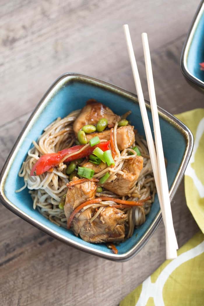 Easy chicken teriyaki recipe with red pepper, carrots, edamame and soba noodles in a small blue bowl with chopsticks on a wooden table.