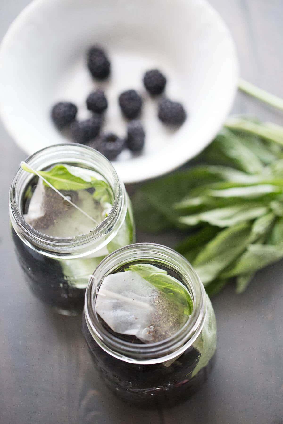 Two glass jars with tea bags and basil leaves next to a white bowl of blackberries on a wooden table.