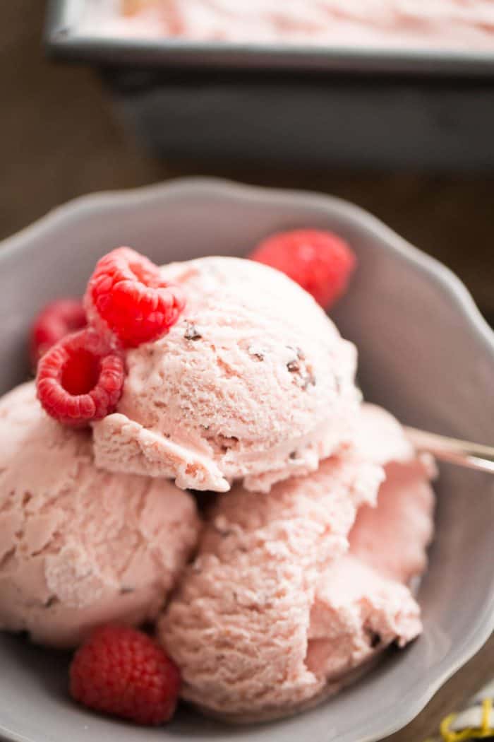 Three scoops of raspberry ice cream topped with raspberries in a small grey bowl with a silver spoon.