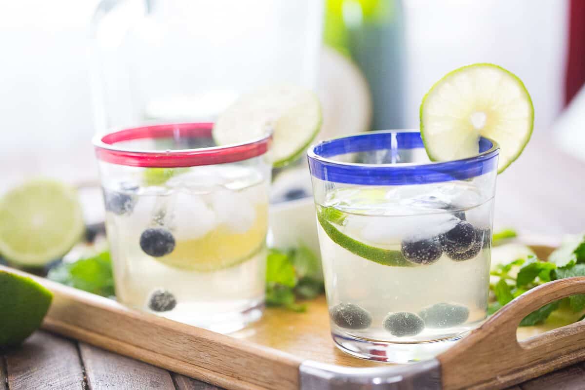Two glasses of sweet blueberry mojitos with sliced limes, blueberries and ice on a wooden platter.