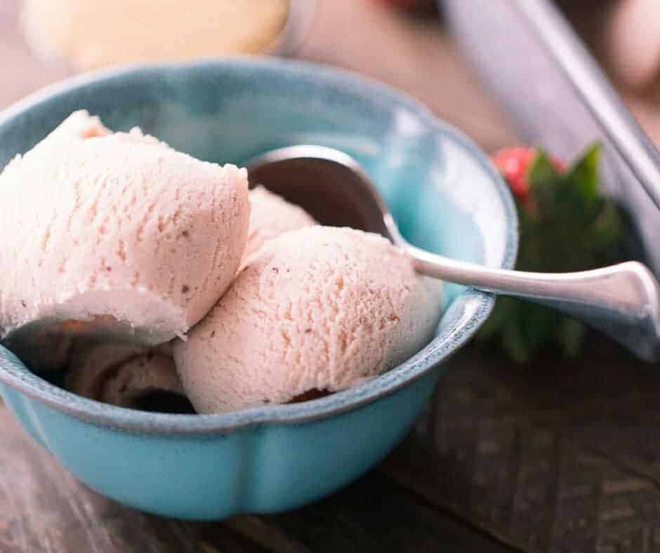 Three scoops of easy Brown Sugar Strawberry Ice Cream and a silver spoon inside of a blue serving bowl on a wooden table.