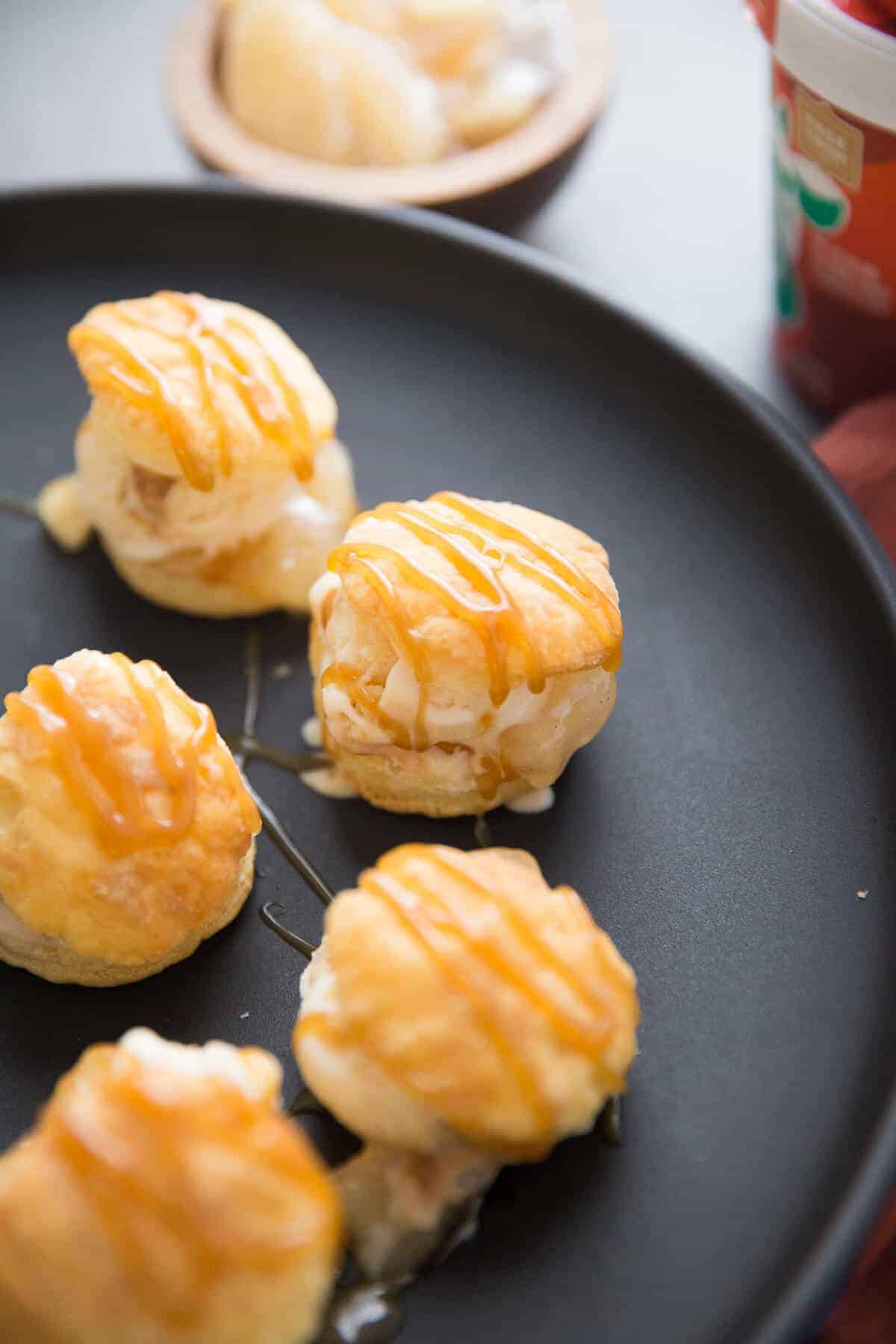 Five easy and delicious profiterole pastries drizzled with caramel on a black serving plate.