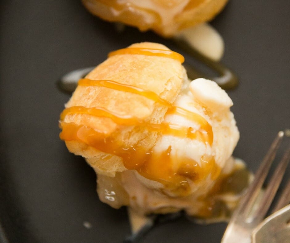 One easy profiterole drizzled with caramel and vanilla ice cream on a black serving plate with a silver fork.