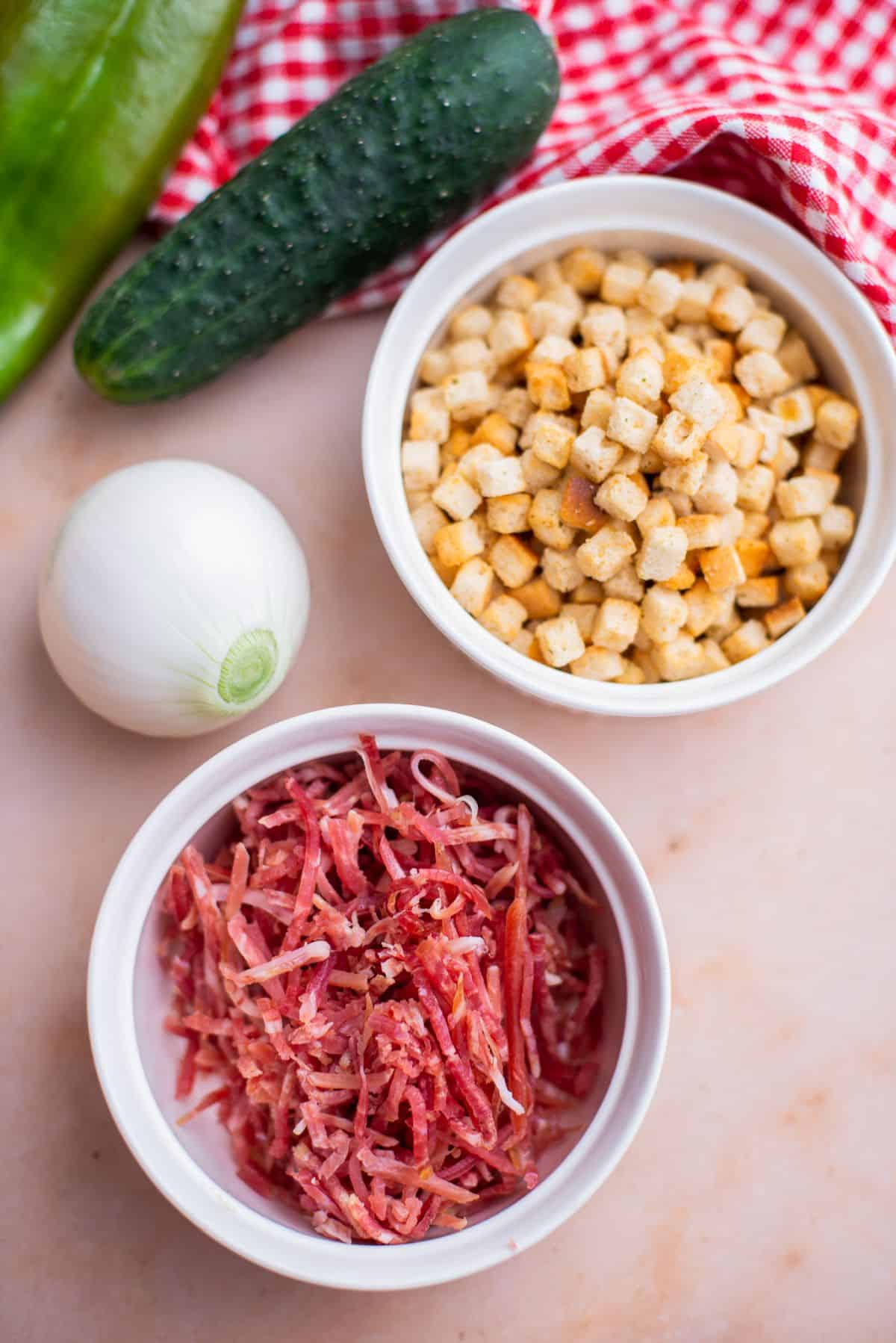 croutons in a white bowl and ham in a white bowl a cucumber and white onion